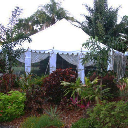 A large white tent with draped fabric siding, nestled among lush green tropical plants and shrubs in a garden setting.