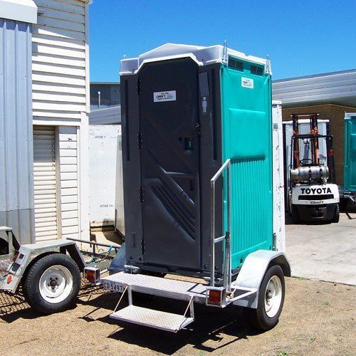 A portable chemical toilet mounted on a small utility trailer outdoors, parked next to a forklift and a storage building.