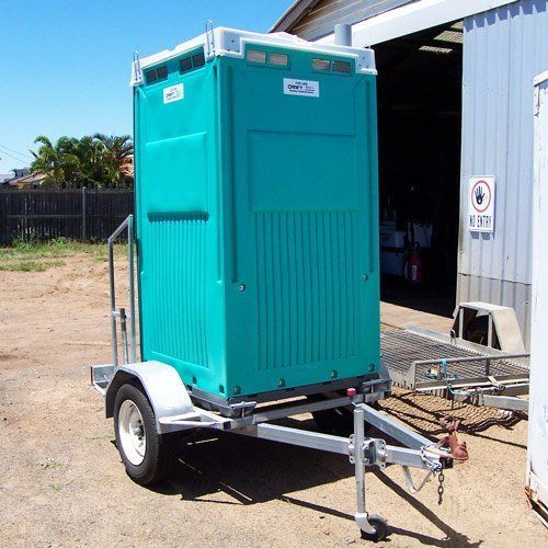 A teal portable toilet mounted on a small metal utility trailer parked outdoors on a dirt lot.