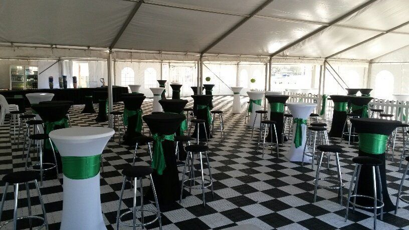 A white marquee tent set up for an event, featuring high-top tables with black and white covers, green sashes, and stools.