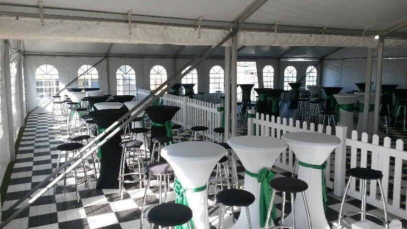 A white party tent interior with high cocktail tables, checkered flooring, and white picket fences.