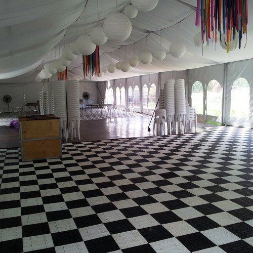 A tent interior with a black-and-white checkered floor, white paper lanterns, colorful streamers, and stacks of chairs.