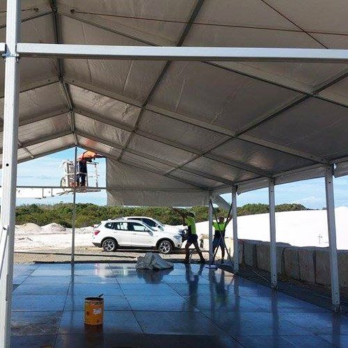 Two workers in high-visibility vests assemble a large tent structure over a concrete floor with a white SUV parked nearby.