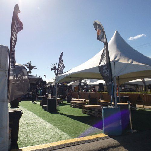 A sunny outdoor beer garden features a white tent, artificial turf, and brand flags set up for an event.