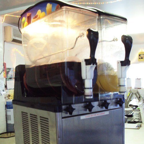 A double-bowl slushy machine sits on a counter, with dark-colored frozen drinks visible in the clear plastic containers.