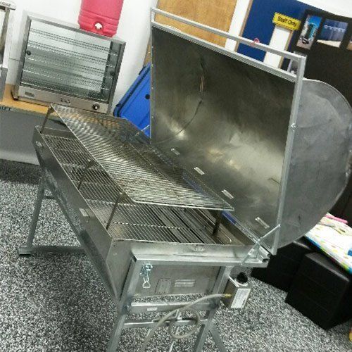 A stainless steel charcoal grill with its lid open, standing on four metal legs in an indoor setting.