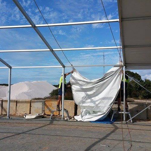 Two workers install a large white fabric roof onto a metal frame structure outdoors near a pile of salt.