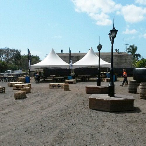 Two white peak tents stand in a gravel area with wooden pallets, straw bales, and street lamps under a sunny blue sky.