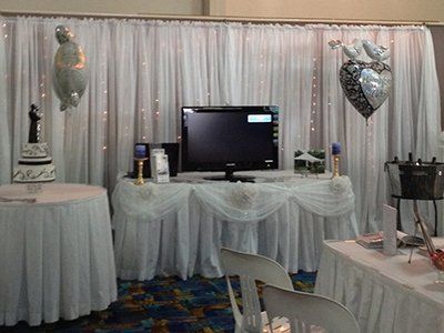 A wedding display featuring tables with white cloths, a tiered cake, a television, and heart-shaped balloons.