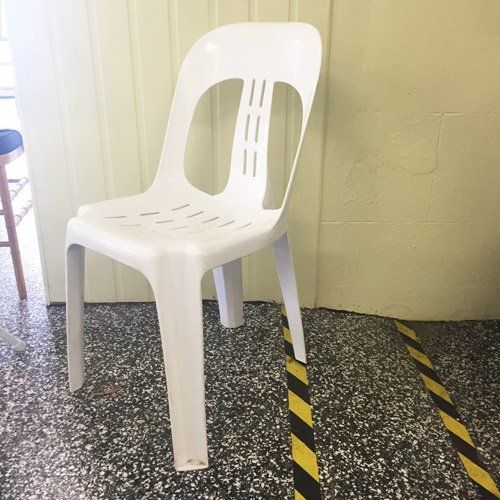 A white plastic chair sits on a speckled floor near a light-colored wall with yellow and black caution tape on the floor.