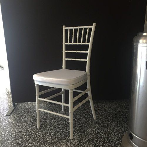 A white chiavari chair with a padded seat stands on a speckled floor against a black backdrop.