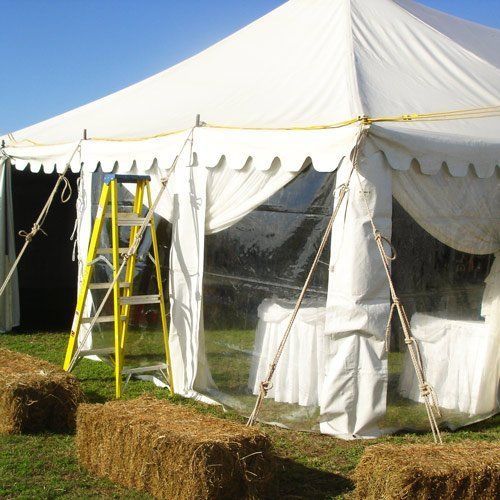 A yellow ladder stands outside a white event tent with clear plastic windows, surrounded by scattered hay bales.