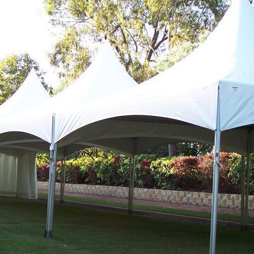 A white, high-peak event tent stands on a grassy lawn in front of a stone wall and leafy green bushes.