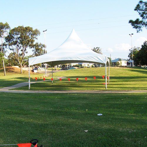A white, peaked event tent stands in a grassy park on a sunny day, surrounded by a line of small orange marker flags.