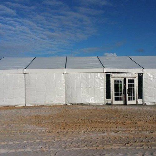 A white temporary event tent with French-style glass double doors stands on a sandy, tire-tracked lot under a blue sky.