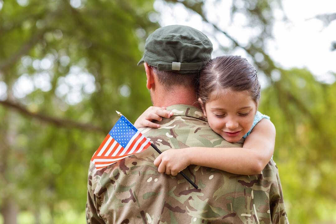 A soldier in camouflage holding a young girl, with an American flag, outdoors. The girl is hugging the soldier.