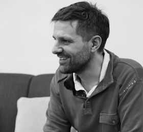Man smiles, looking to the side. Wearing a long-sleeved shirt with buttons and a collared undershirt. Seated indoors.