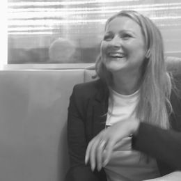 Woman laughing, wearing a blazer over a t-shirt, seated indoors.