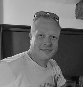 Man with sunglasses on head, smiling. Wearing white t-shirt. Interior setting with dark background.