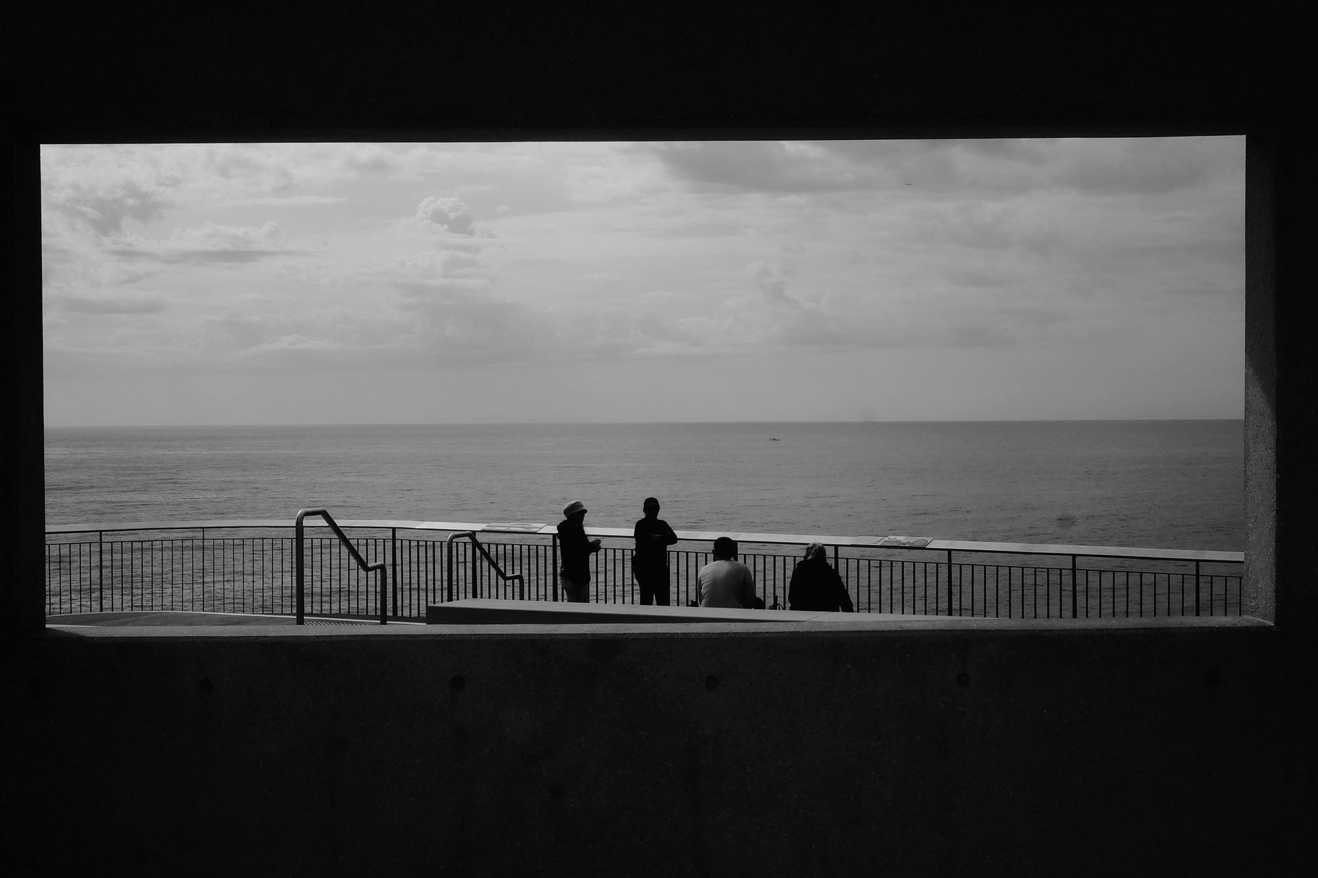 Silhouetted people on a seaside walkway, gazing at the ocean through a rectangular opening.