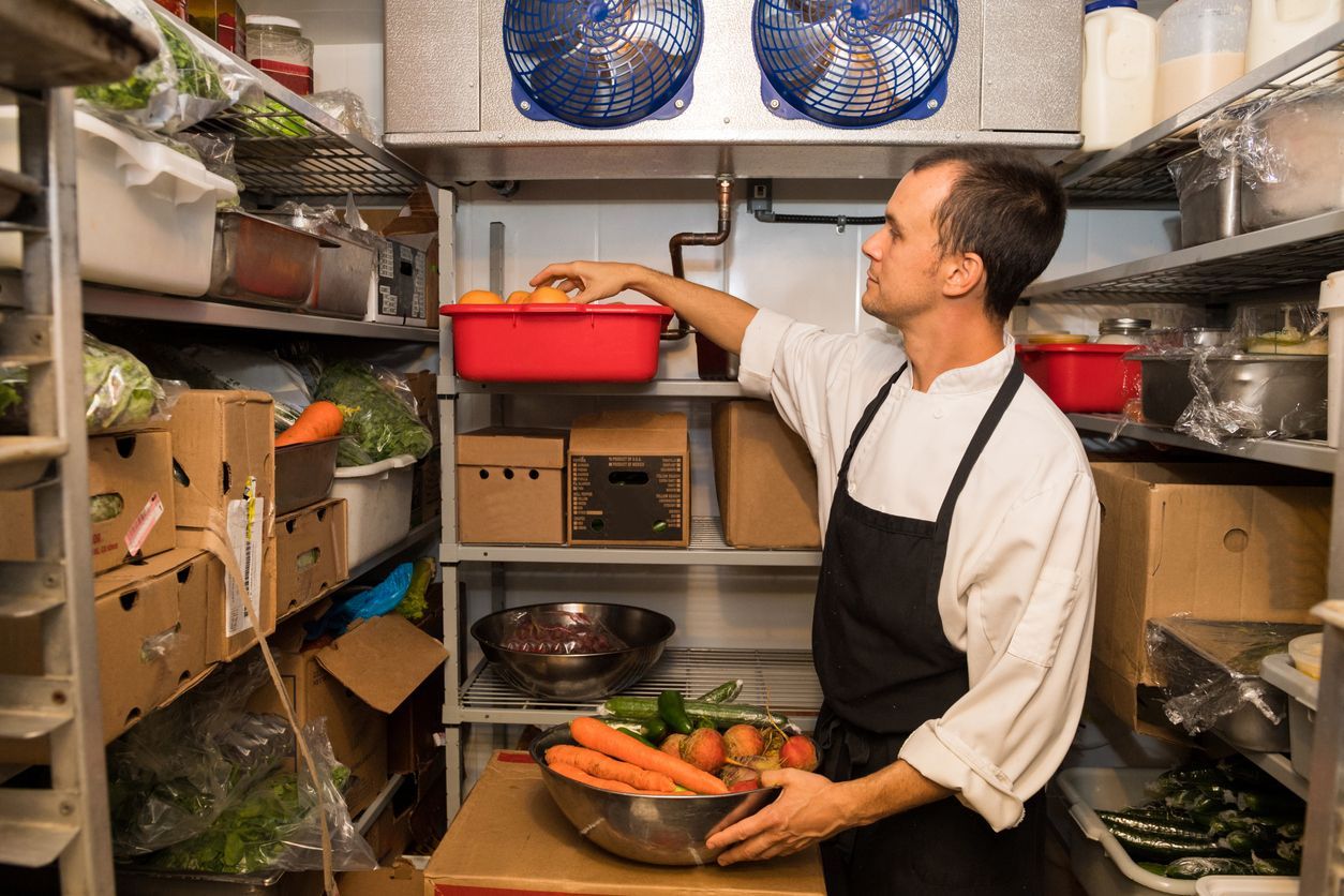 Person stocking produce in a small storage room, reaching for a red bin on a shelf.
