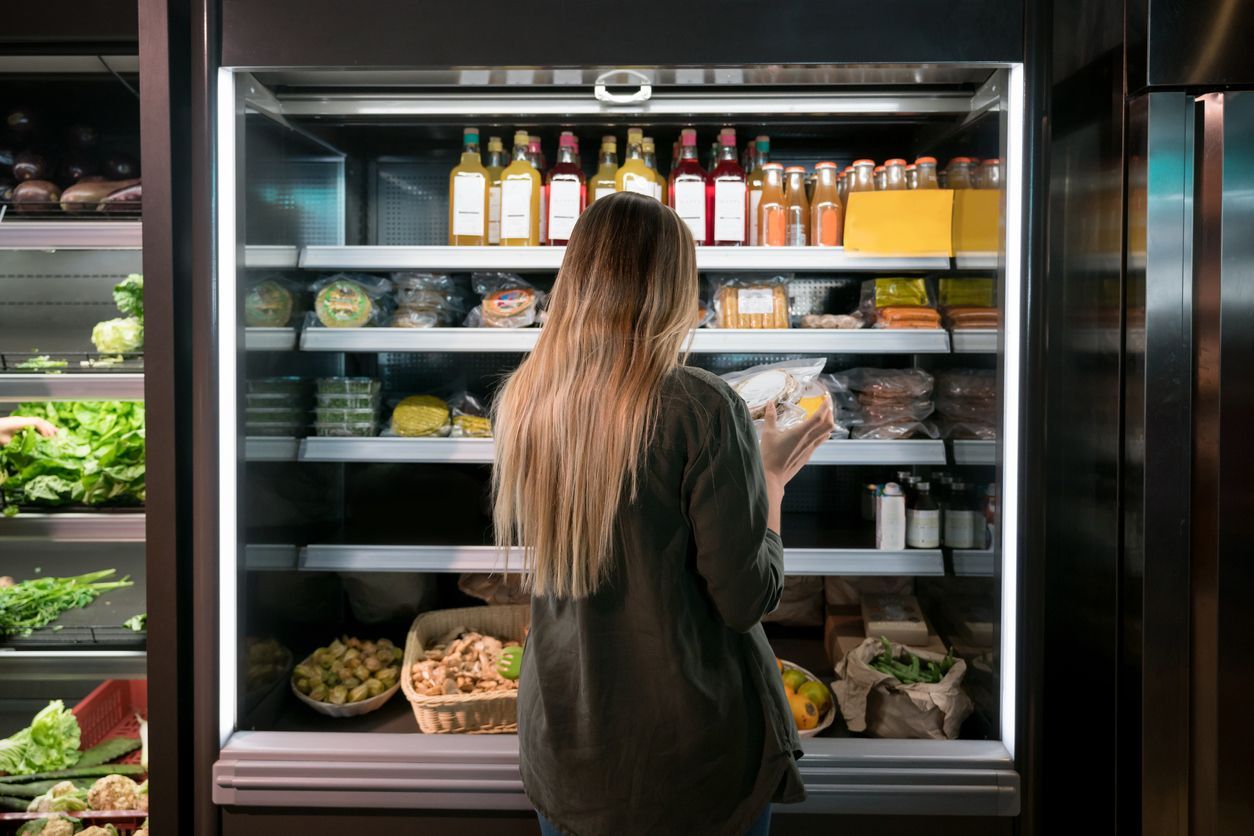 Woman browsing items in a brightly lit grocery freezer case with shelves of food and drinks
