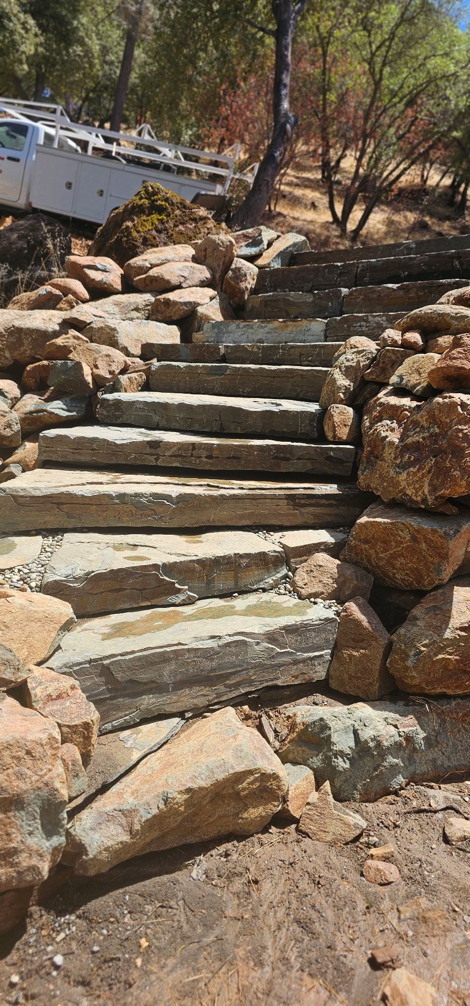 Rocky outdoor stone steps winding up a wooded hillside with a white wall on the left