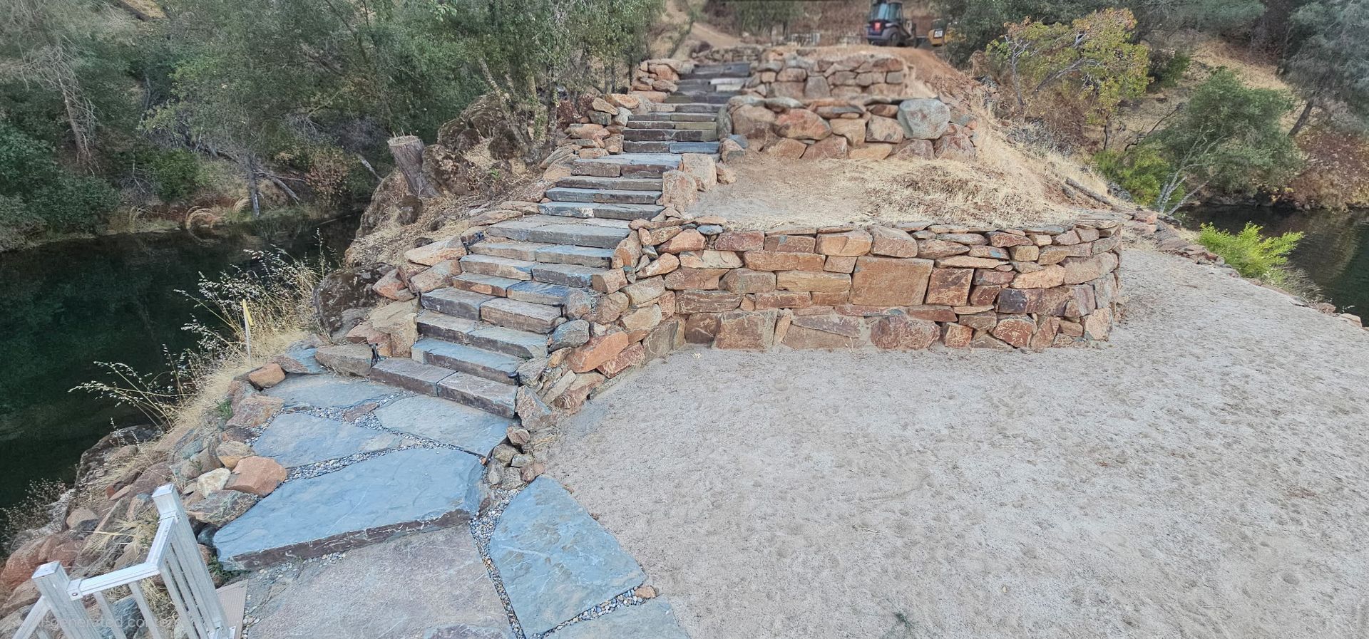 Stone steps built into a rocky hillside beside a gravel path and creek, surrounded by shrubs.