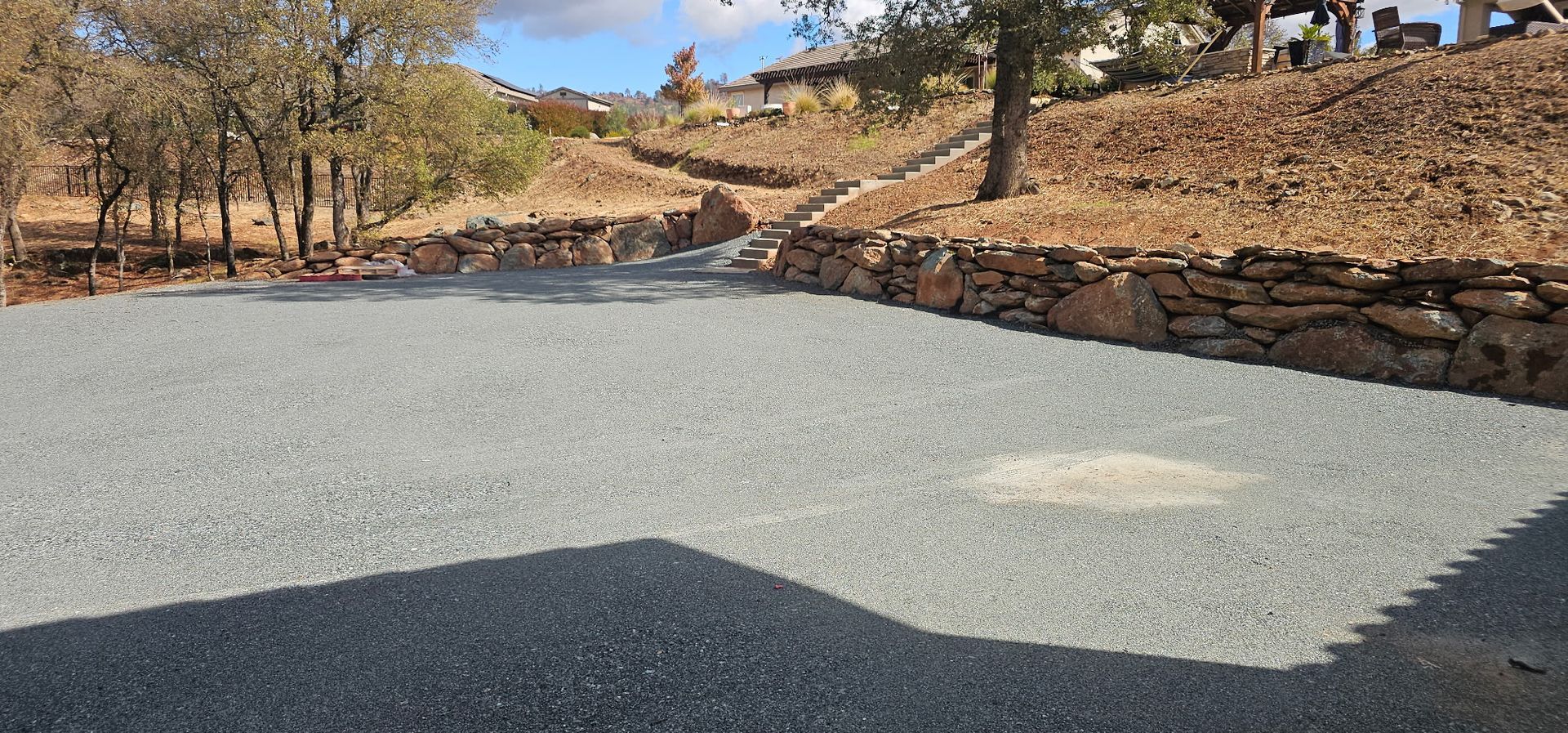 Gravel driveway beside low stone walls in a dry, hilly landscape with sparse trees and blue sky