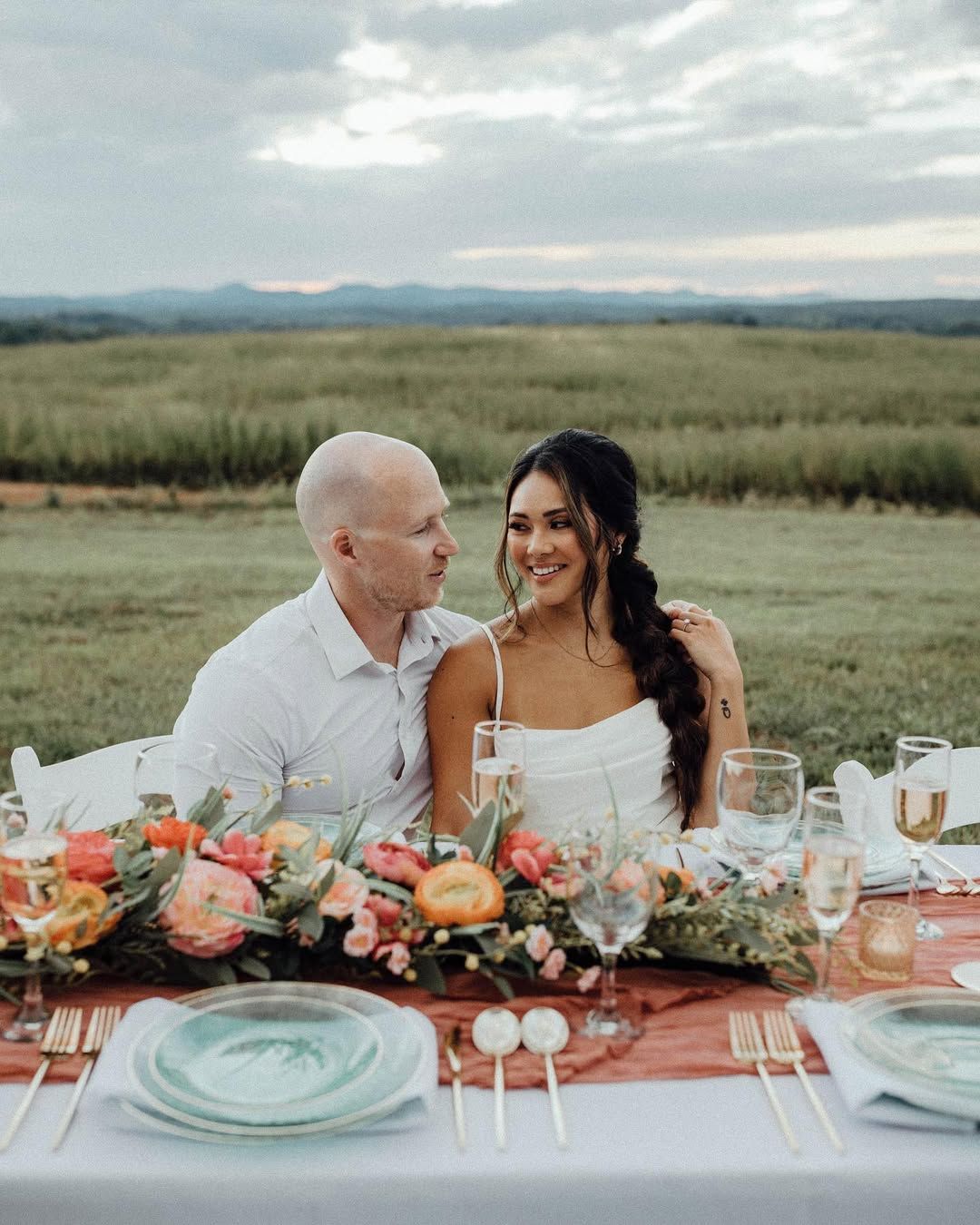 Couple at a table outdoors, smiling at each other. Floral centerpiece, gold flatware, and scenic field background.