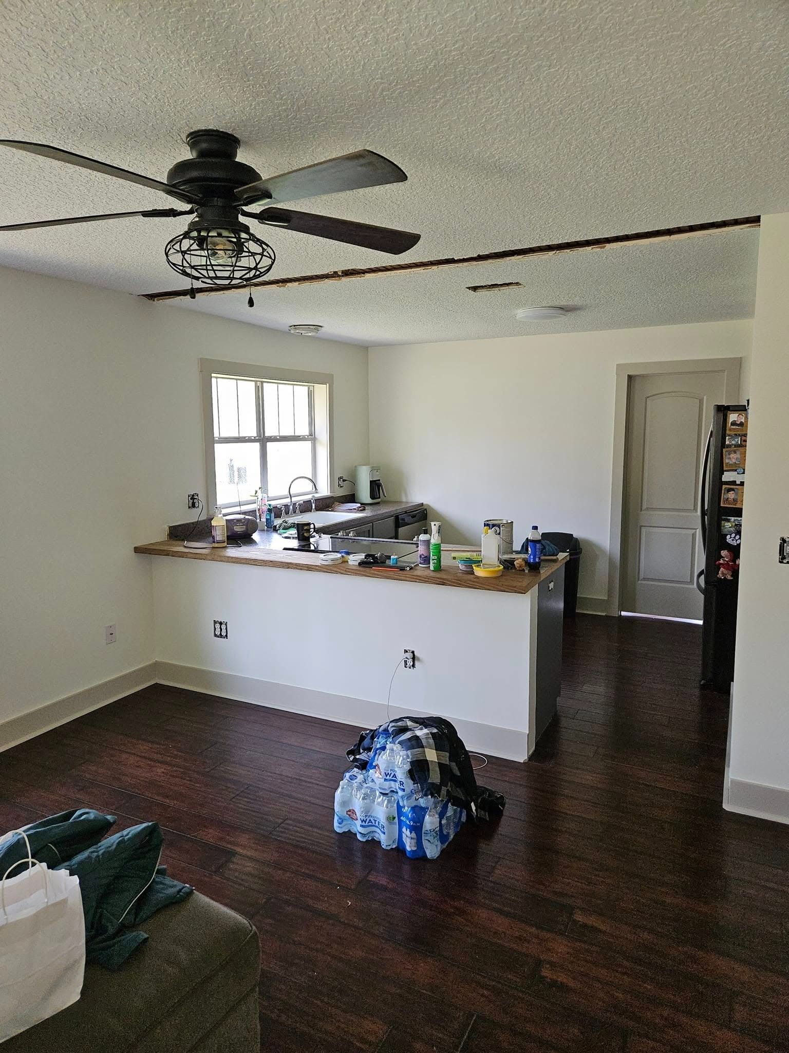 Living room with dark wood floors, ceiling fan, open kitchen counter, and a backpack on the floor