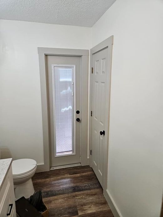 Small bathroom with white walls, dark wood floor, toilet, and narrow glass-paneled door with closet beside it