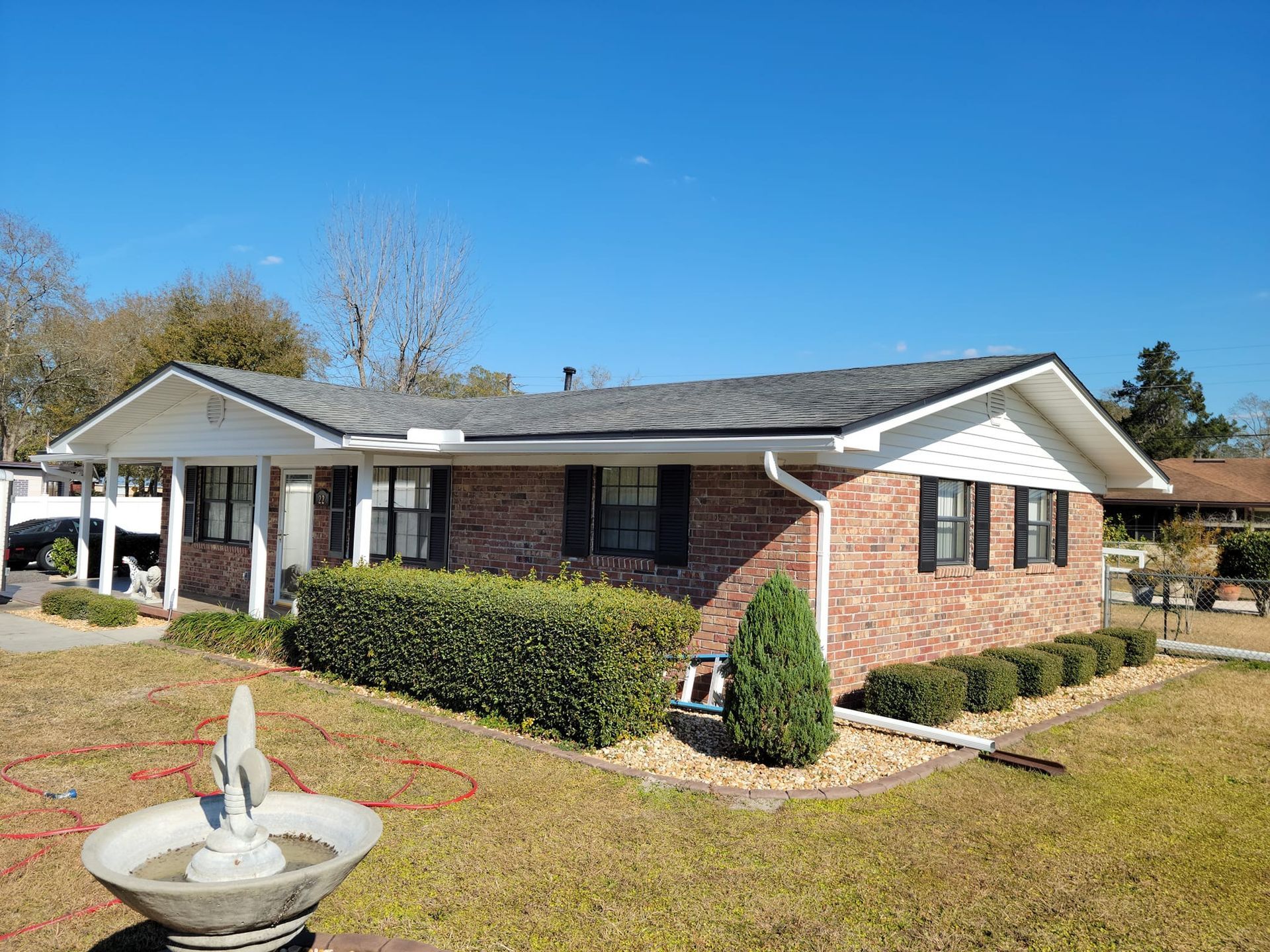 Brick ranch house with white trim, black shutters, and a small fountain in the front yard under a blue sky