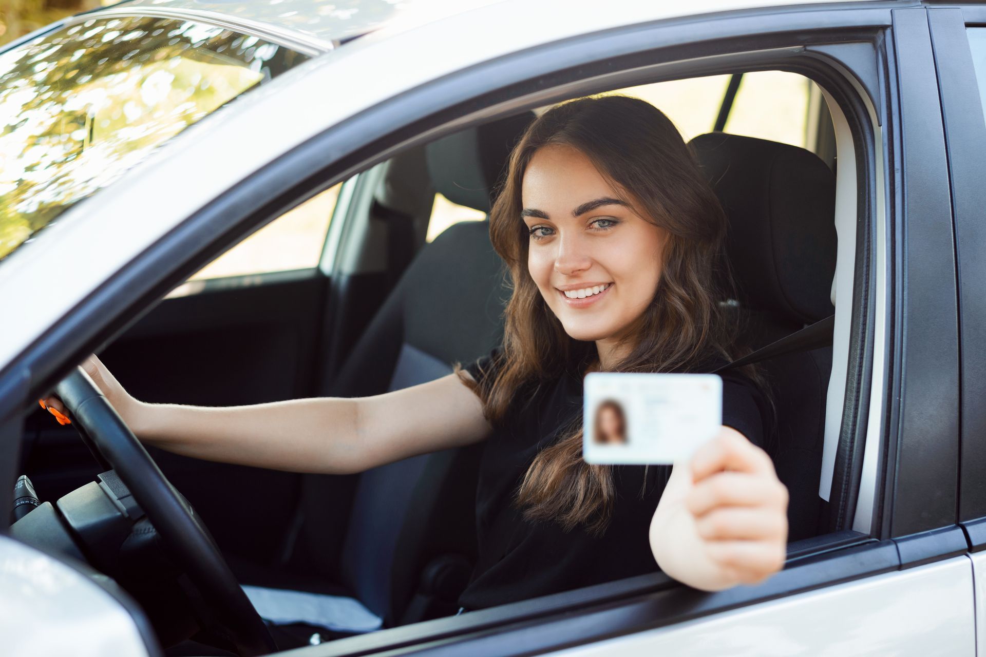 A happy student driver is seated in a modern silver car while showing her driver's license.