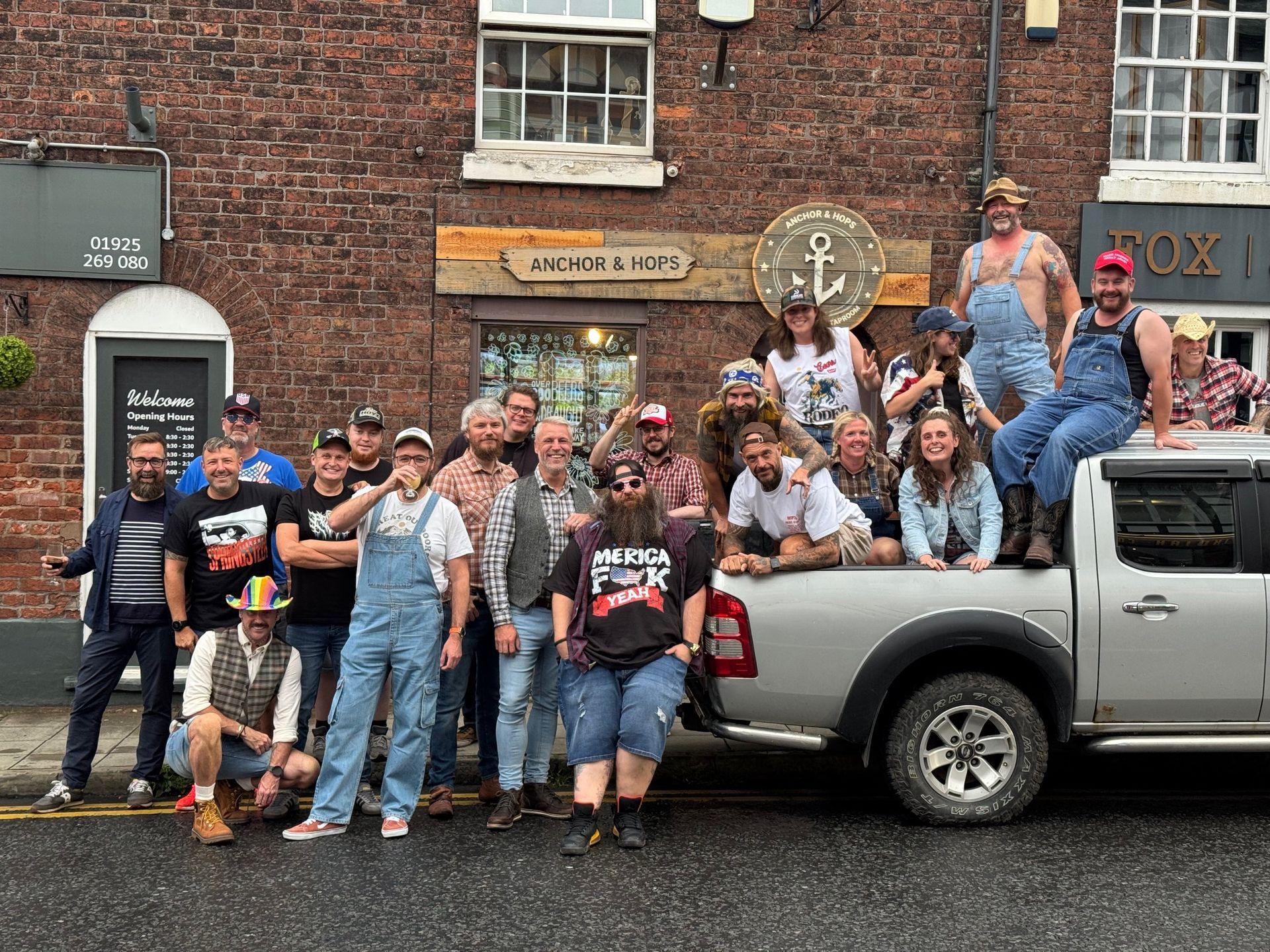 A large group posing outdoors in front of a brick building and a silver pickup truck, some people sitting in the truck bed.