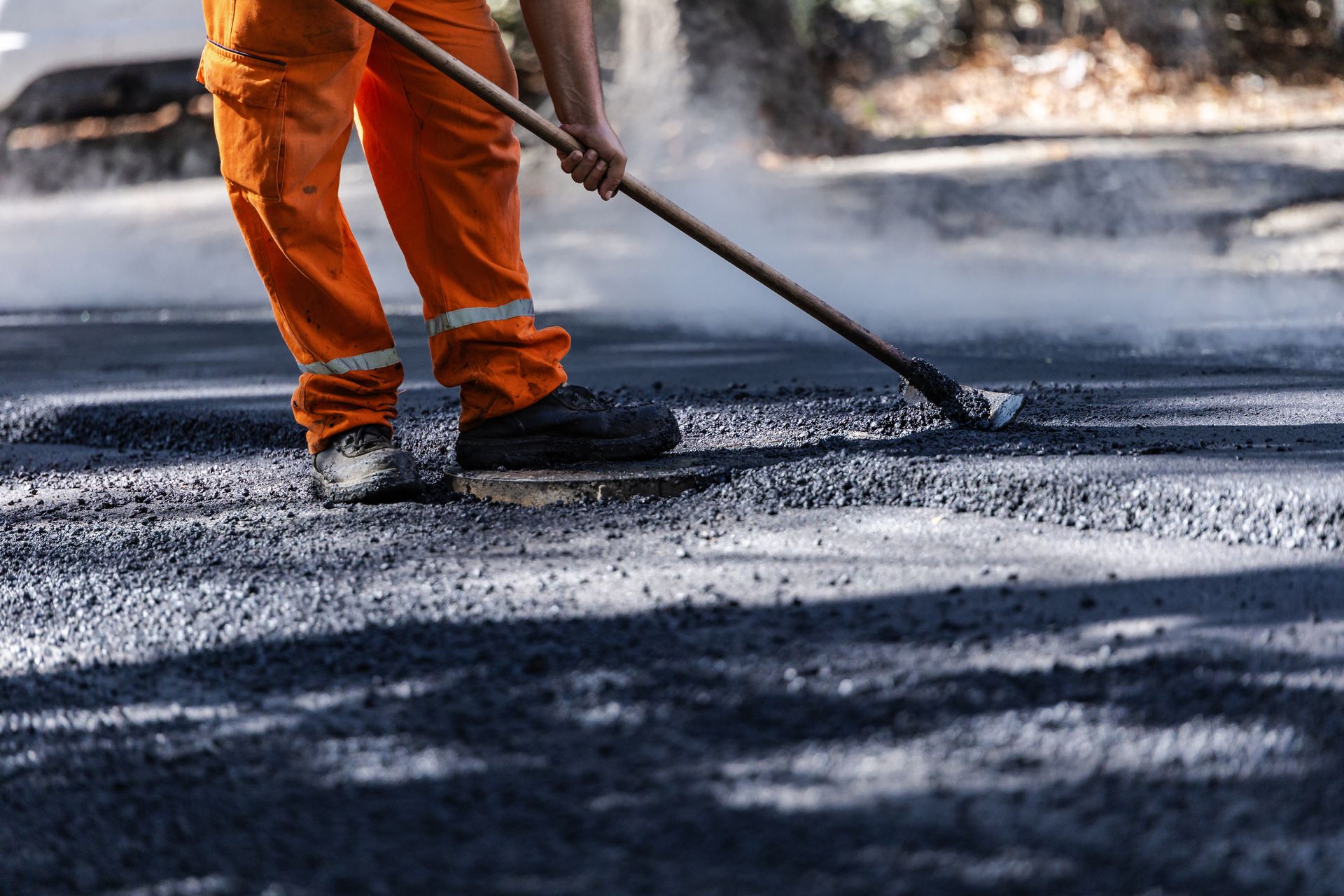 Road worker in high-visibility clothing spreading and leveling hot asphalt during road paving.