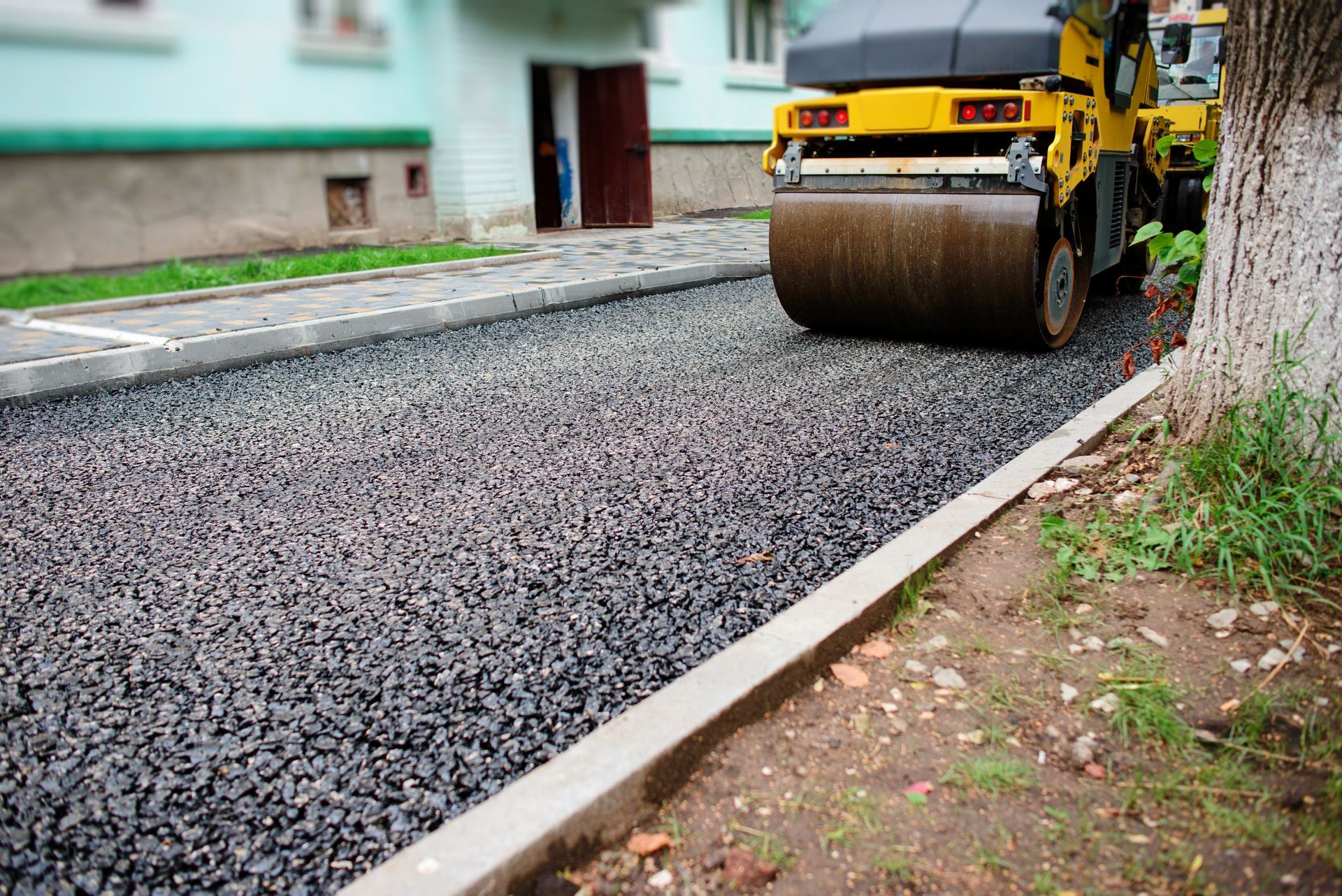 Yellow road roller compacting fresh asphalt on a residential street.