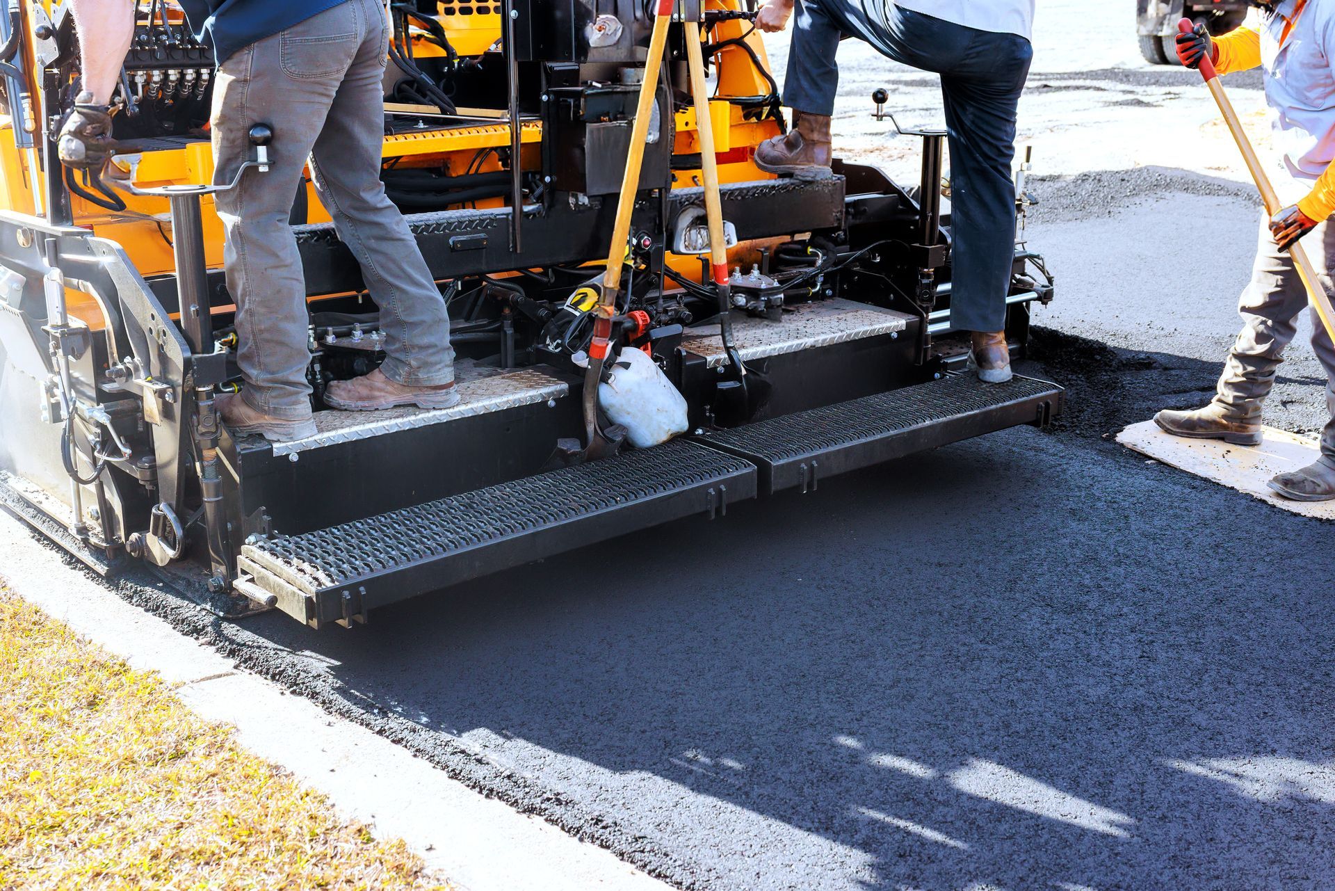 Road crew operates asphalt paving machine while laying fresh pavement on street.