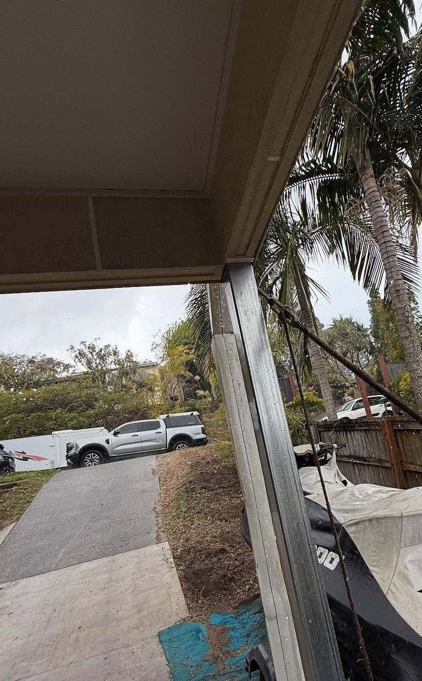 A galvanized steel post supports the corner of an outdoor roof overhang, with a blurred driveway and trees in the distance — RAC Carpentry Services in Pimpama, QLD