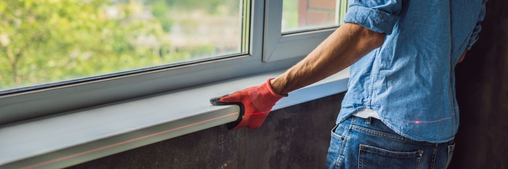 A person in a blue shirt and red work glove installs a white windowsill beneath a bright window — RAC Carpentry Services in Oxenford, QLD