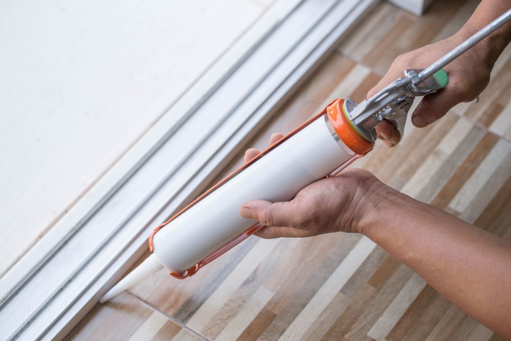 A person using a caulk gun to apply sealant along the edge of a tiled floor near a door frame — RAC Carpentry Services in Coomera, QLD