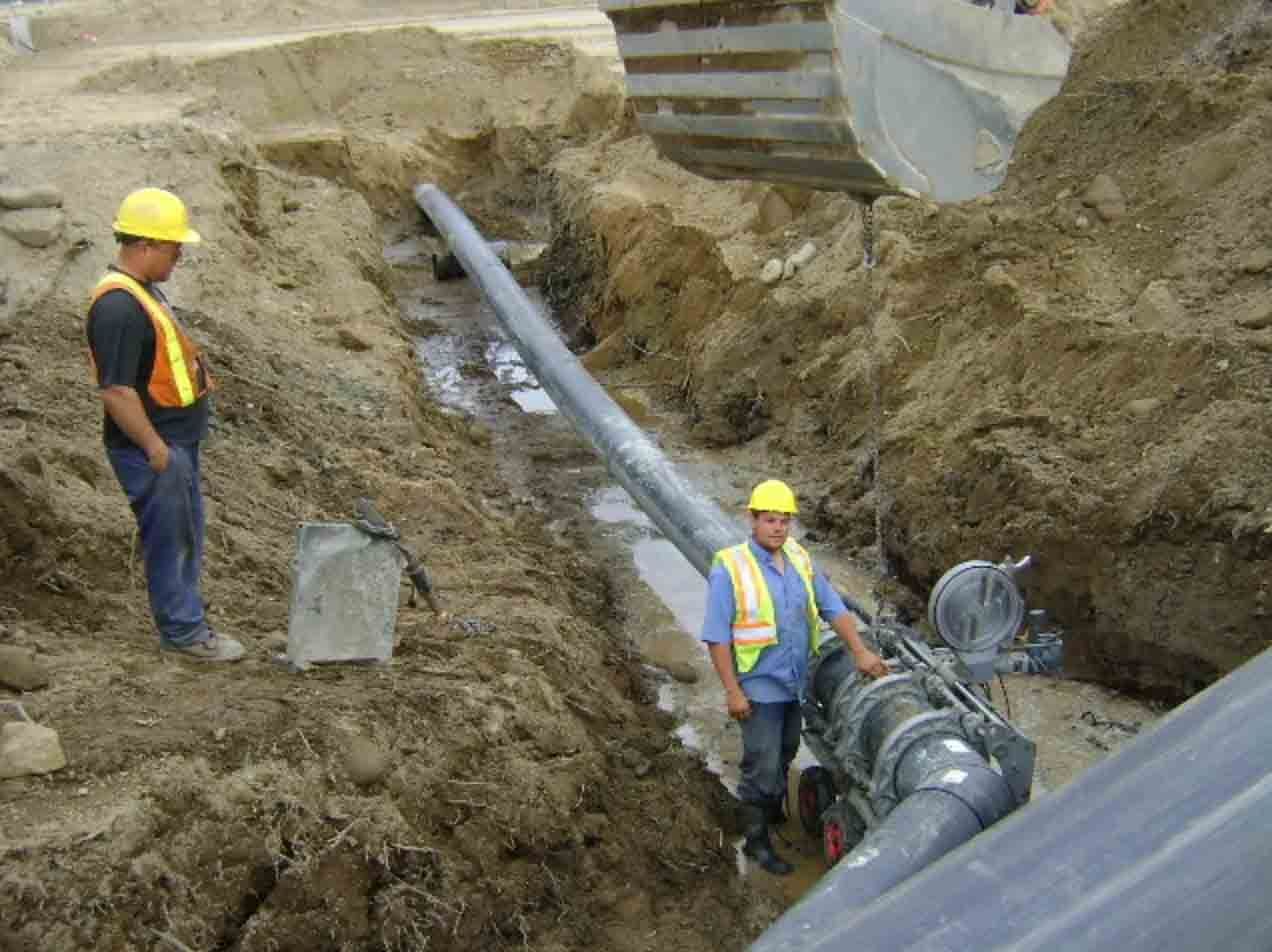 Des ouvriers du bâtiment, casqués, installent de grosses canalisations souterraines dans une tranchée.