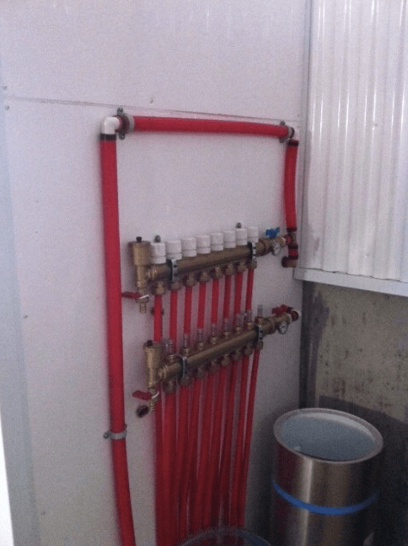 Collecteur de plomberie rouge avec plusieurs tuyaux et vannes, fixé au mur à côté d'un seau en métal.