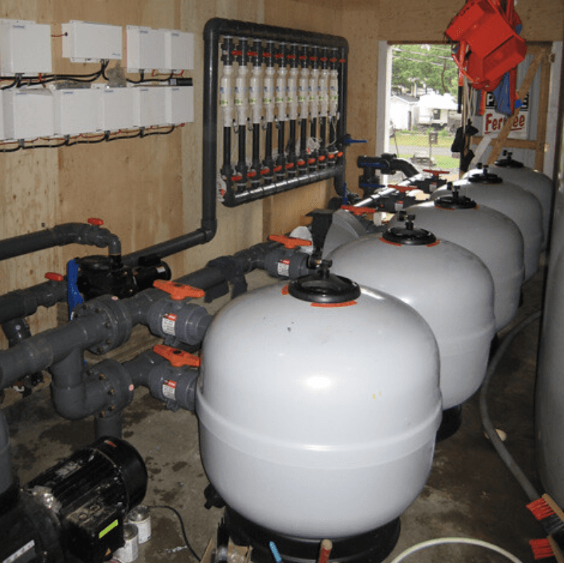Rangée de grands réservoirs de filtration blancs et de tuyaux dans un local technique, avec un panneau de commande au mur.