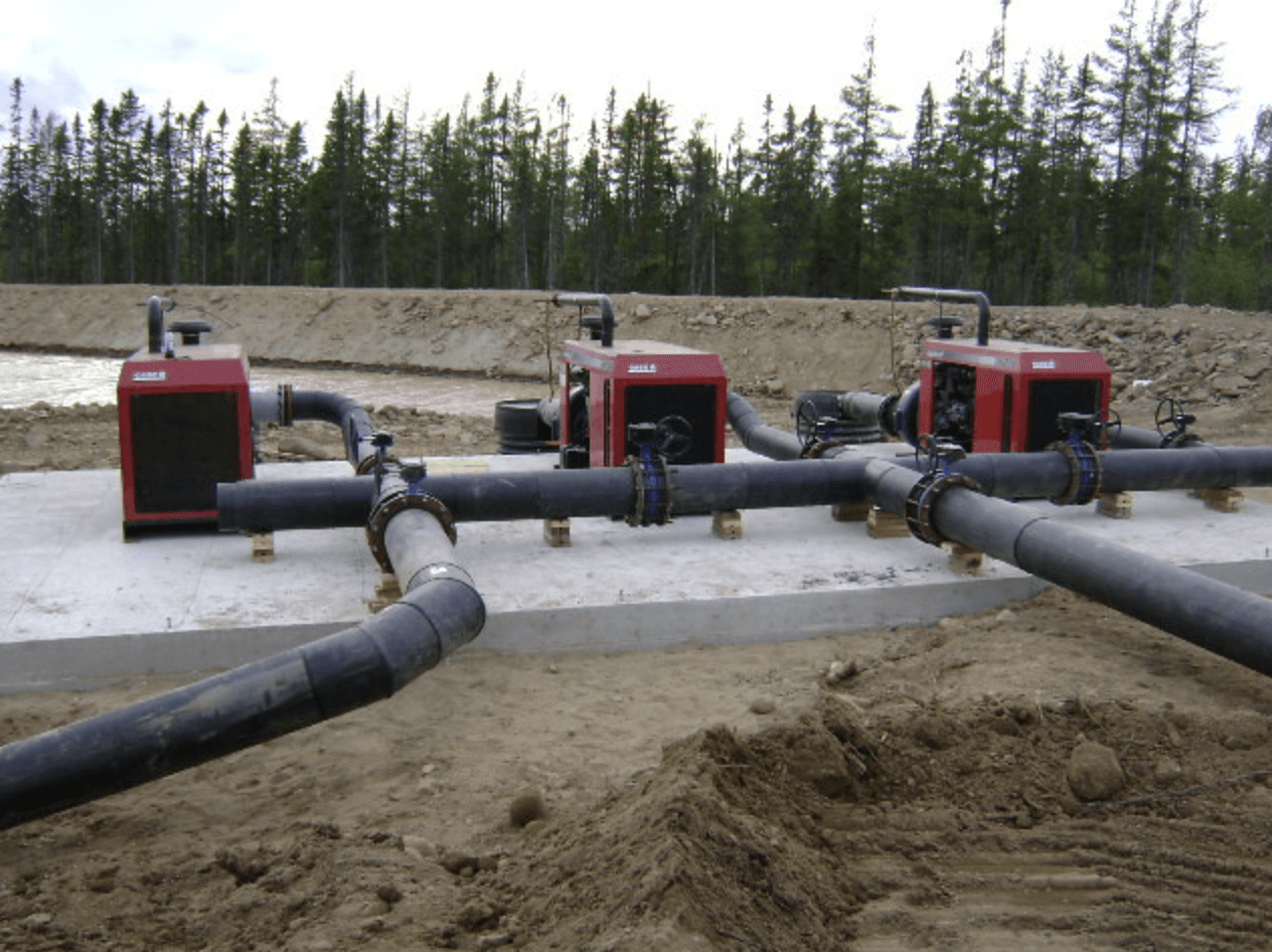 Trois pompes industrielles rouges reliées à de gros tuyaux noirs sur un chantier boueux avec des arbres en arrière-plan