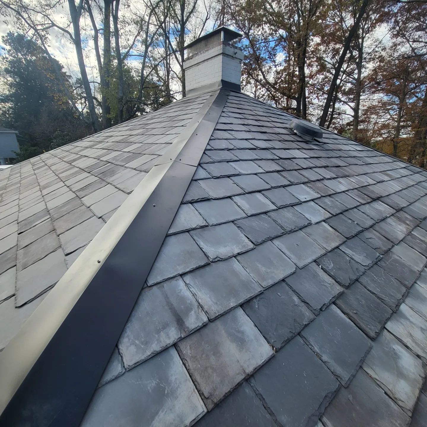 Roofer on a gray shingled roof, hammering shingles. Sunny day, house in background.