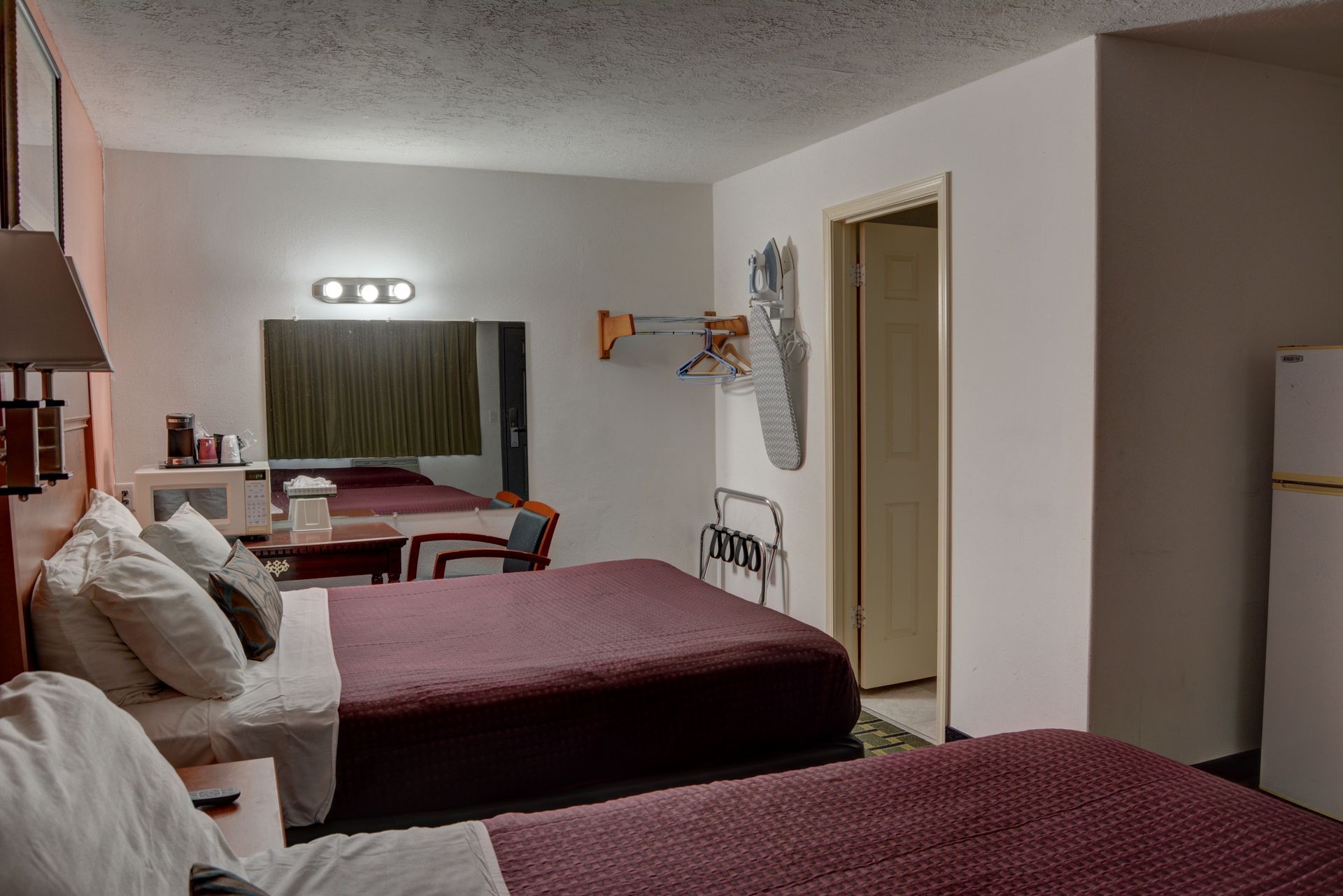 A motel room with two beds, a mirror, and a refrigerator. The walls are white, and the beds have maroon covers.