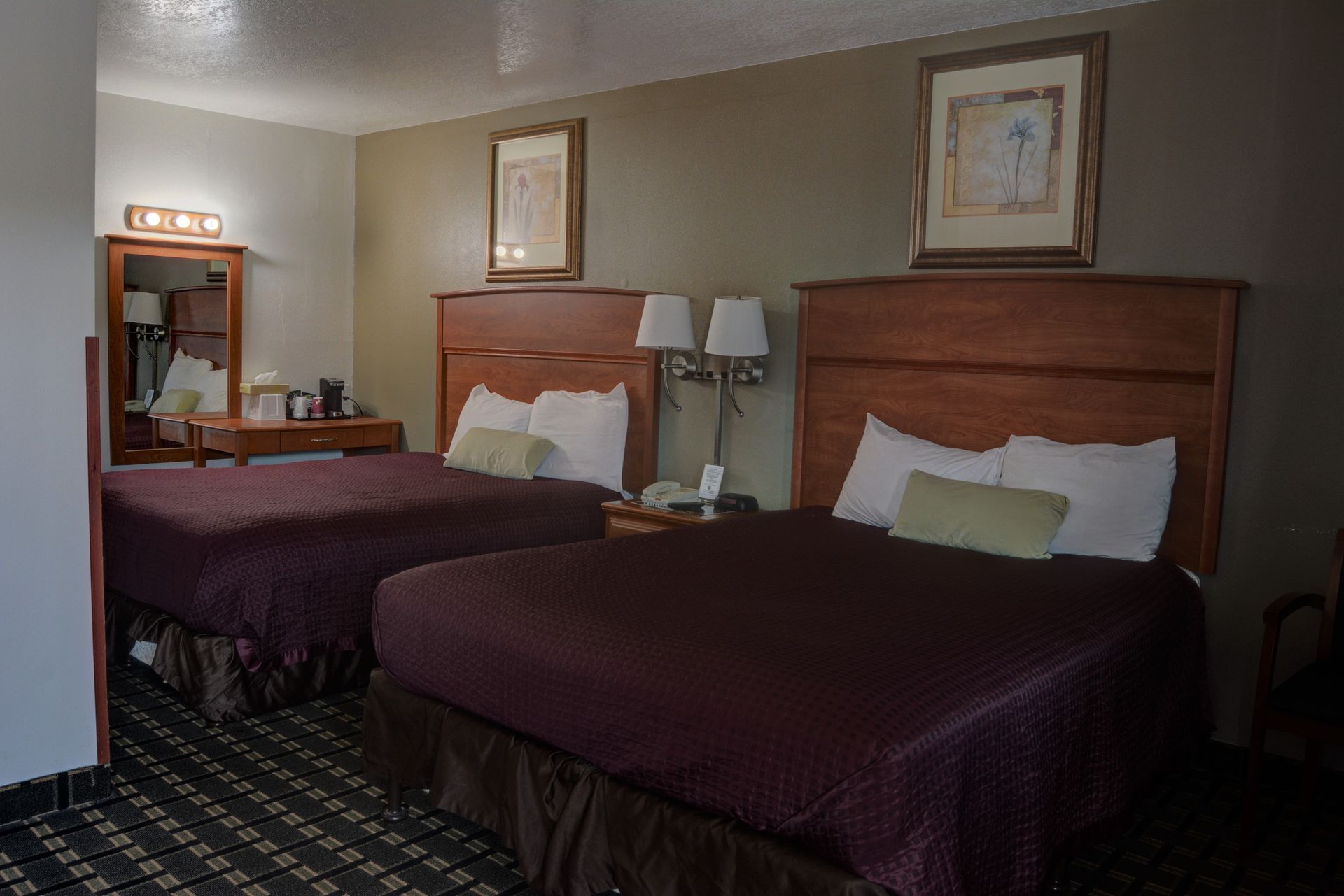 A hotel room with two double beds, brown headboards, and burgundy bedspreads.  Neutral-toned walls, a vanity