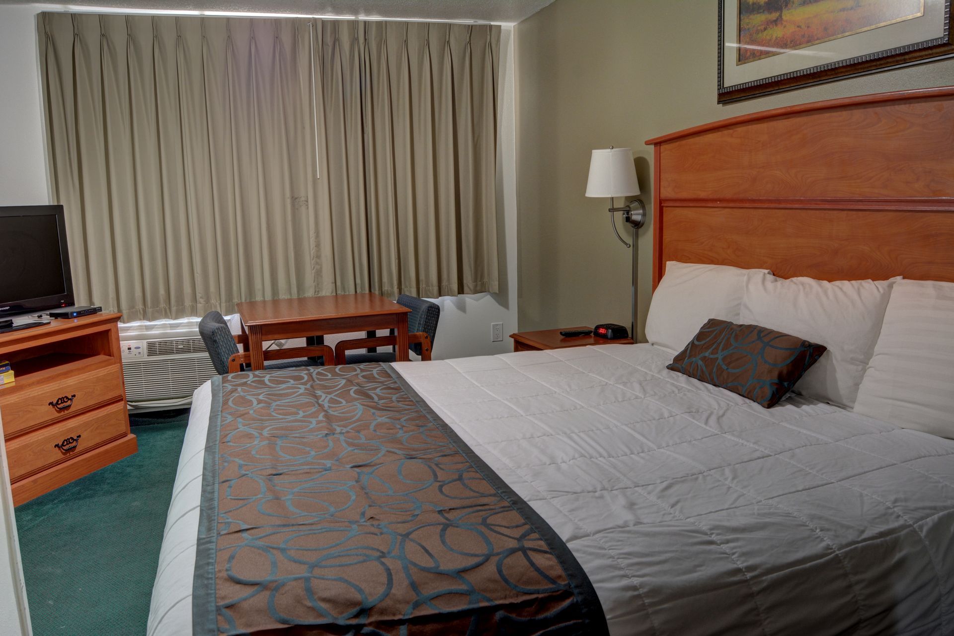 Hotel room interior with a bed, dresser, table, and window.  A television sits atop the dresser. 