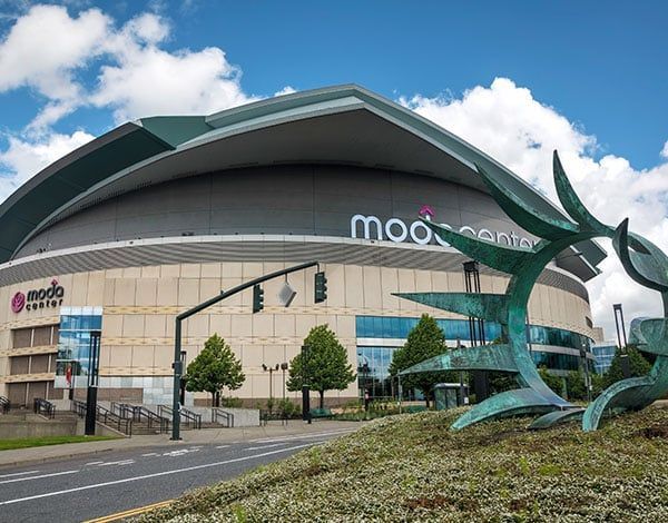 Moda Center arena with a bronze sculpture in the foreground, a blue sky, and a street and sidewalk.